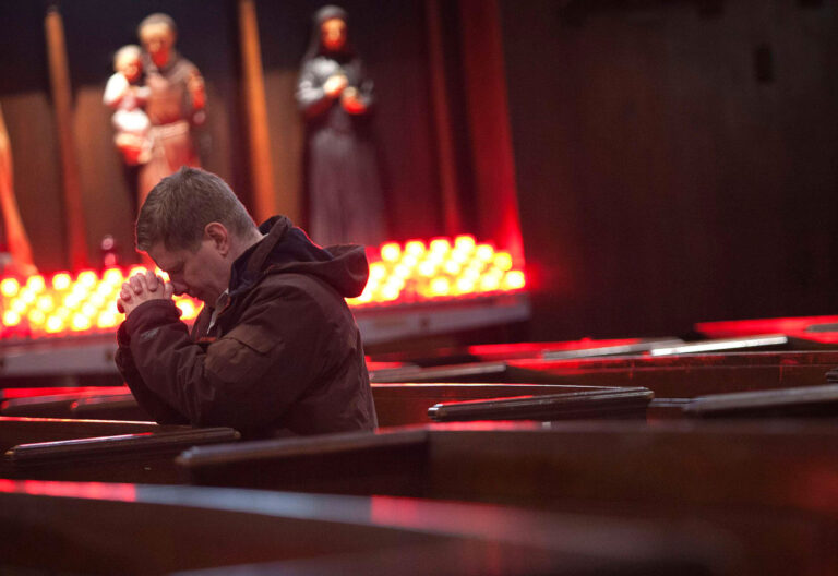 Un hombre reza de rodillas en una iglesia vacía (de fondo, varias imágenes de santos y un lampadario)