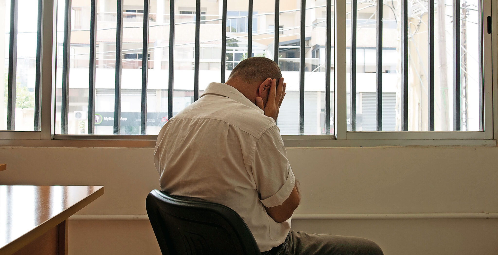 Hombre sentado de espaldas frente a una ventana con rejas y las manos en el rostro